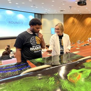 A student and an adult listen to a professor in a white coat while she points to bright green, blue, and yellow images printed on large pieces of shiny paper laying on desks.