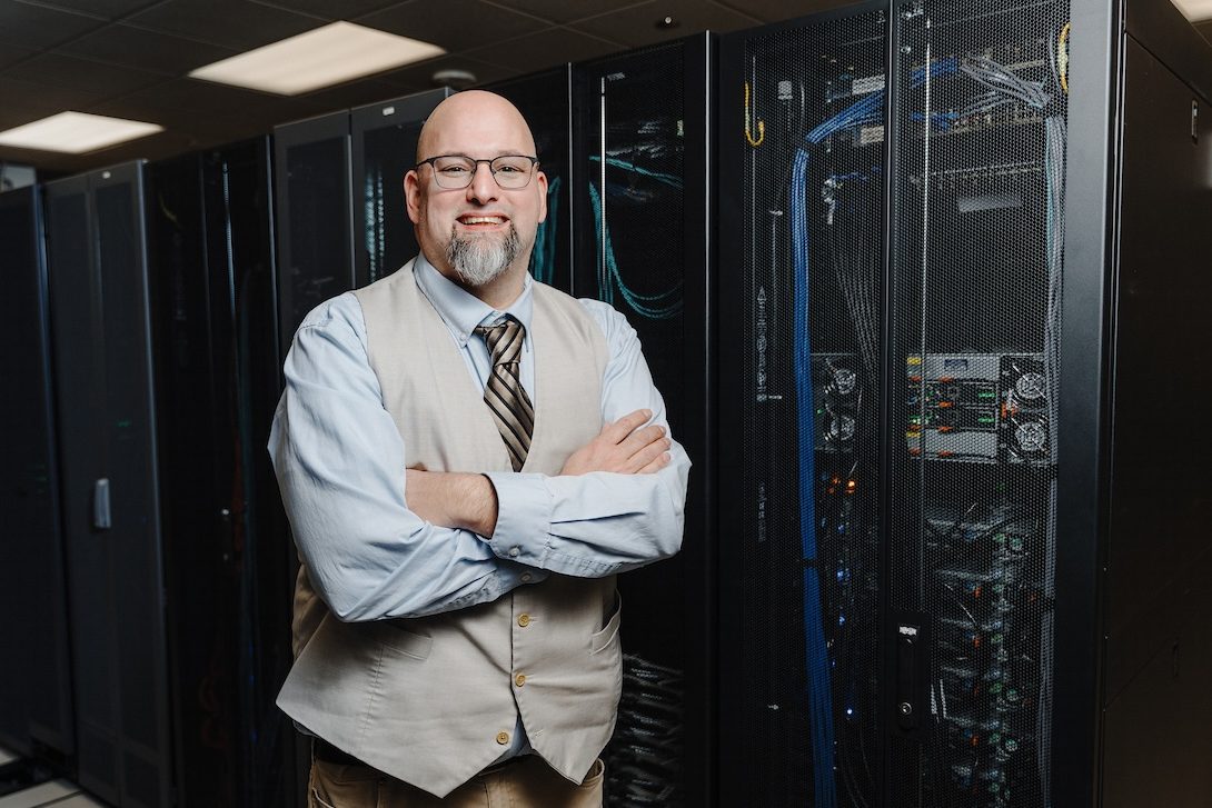 Ammar Ammar stands in front of a row of university servers, with one server's blue chords visible through the mesh covering