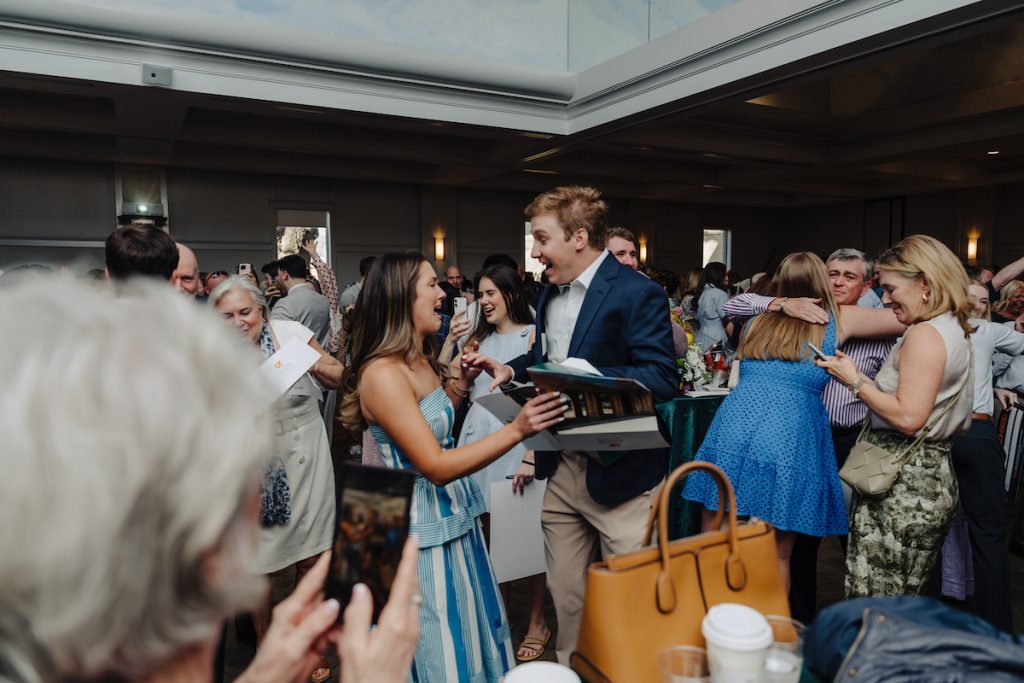 A young woman expresses excitement and tears, celebrating with a young man who's also expressing surprise and excitement with her, while other students embrace loved ones around them.