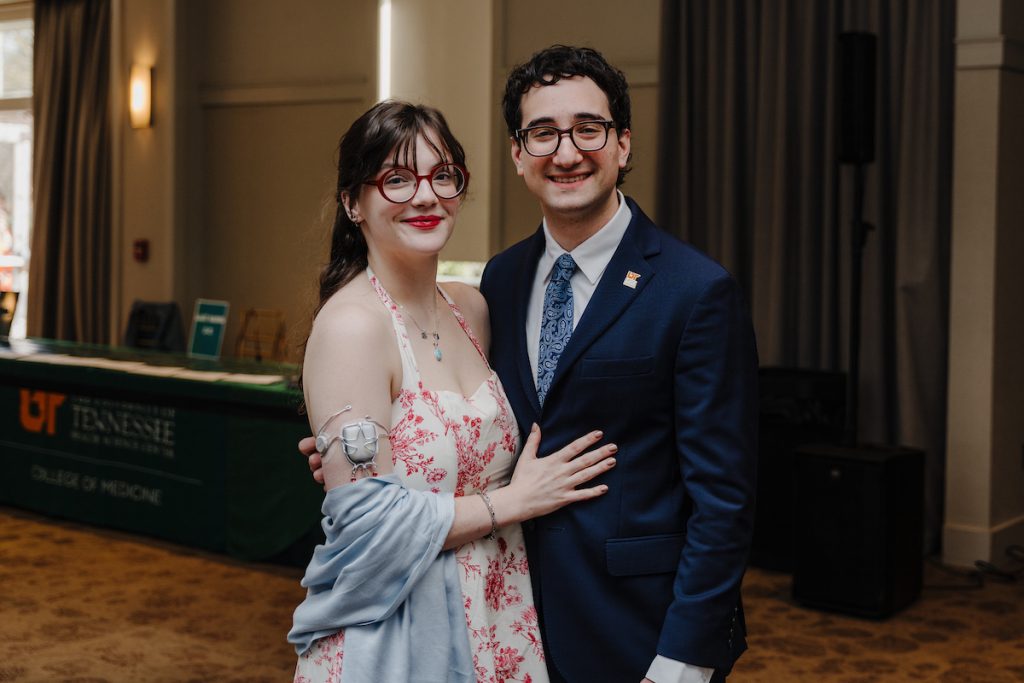 Medical student Ben Finder in a suit with a UTHSC pin and a young woman in a flowered dress pose for a photo indoors. She has a medical device on her arm.