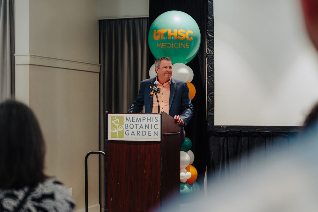 Dean Michael Hocker speaks at a podium with a sign on it that says Memphis Botanic Garden. A pillar of balloons is behind him with a large, green ballon on top that says UTHSC Medicine in orange.
