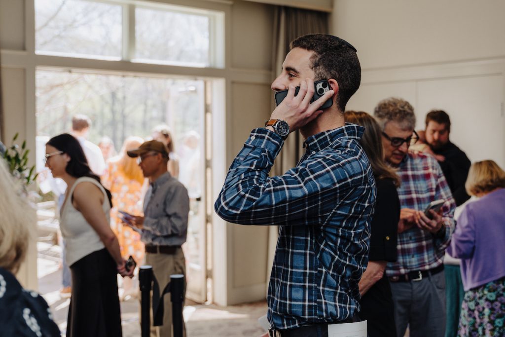 A student in a plaid shirt is holding a cell phone to his ear to discuss the news. Open glass doors are in the background with people inside and outside.