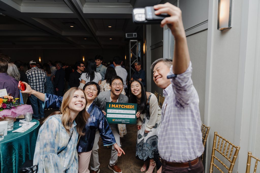 A man holds a cell phone out and up to take a photo of the moment, with a young man holding his "I Matched!" sign alongside family and friends smiling broadly and laughing.