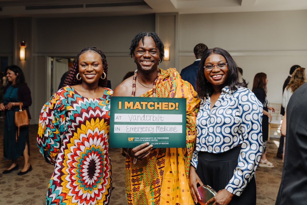 A Black man in African clothing and two Black women in brightly colored and bold print clothing pose for a photo. The man is holding his "I Matched!" sign that reads "at Vanderbilt in Emergency Medicine."
