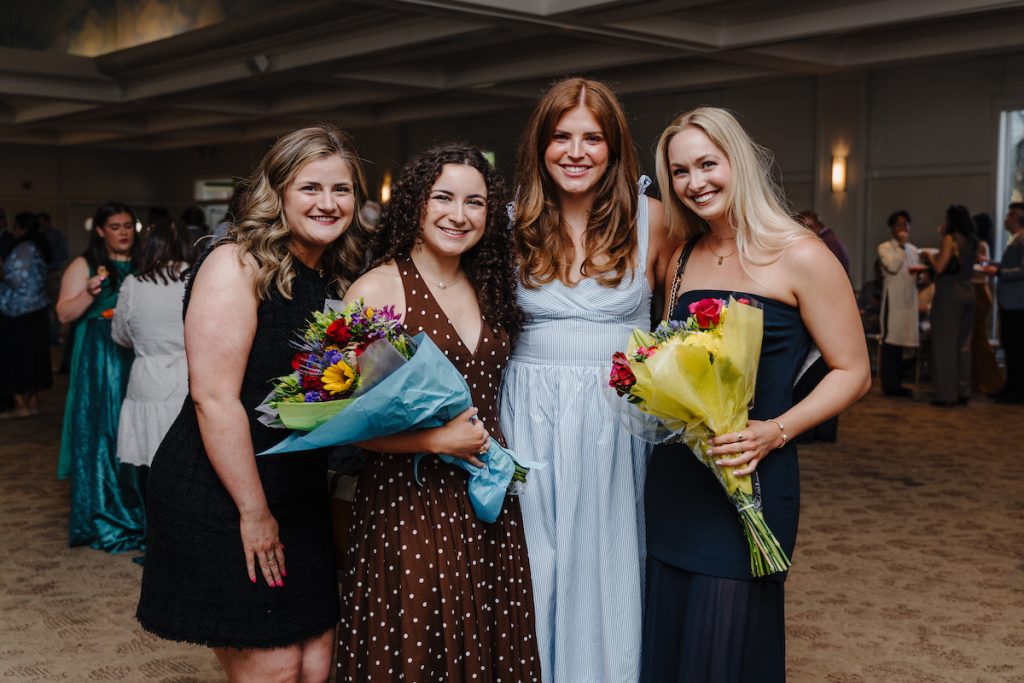A group of four ladies in dresses pose for a photo, two holding bouquets of flowers.