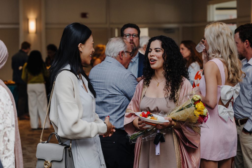 A woman with a bouquet of flowers and plate of fruit talks candidly to another woman who's smiling big.