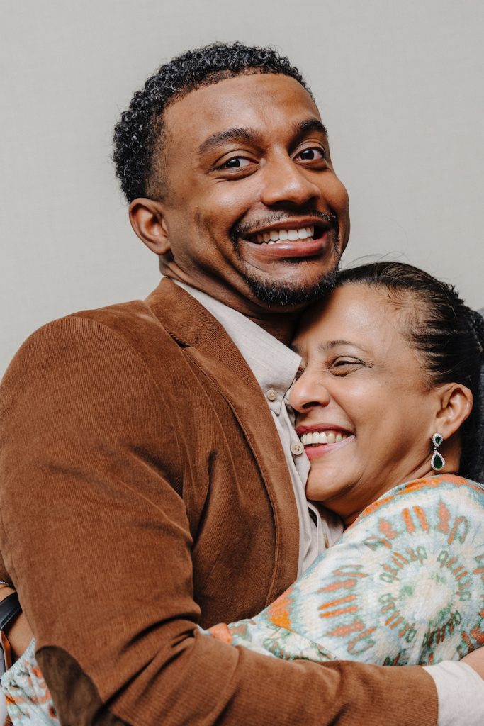 A close-up of a Black male student smiling big and being hugged tightly by a Black woman.