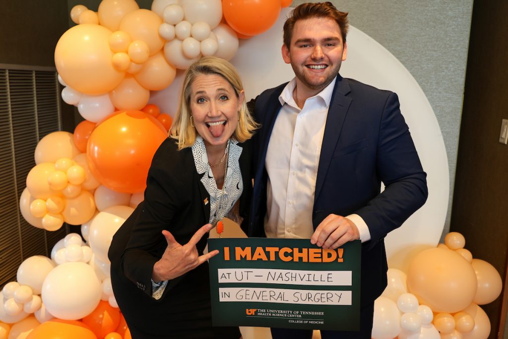 In Knoxville, a student poses with an older woman in front of orange balloons. She has her tongue out while she smiles and is making an "I love you" symbol with her fingers. She has a UTHSC pin on her lapel. His "I Matched!" sign reads "at UT – Nashville in General Surgery."