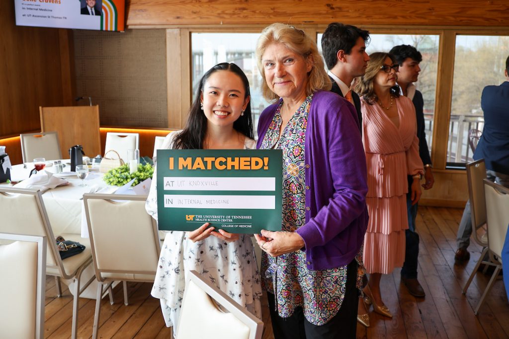 A young Asian female student poses with an older white woman in front of a set table and some others as well as a wall of windows, in Knoxville. Her "I Matched!" sign reads "at UT – Knoxville in Internal Medicine."