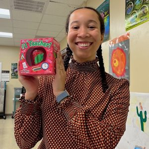 In a high school classroom, Hannah holds up a gift of a toy apple that says #1 teacher on the box, with science posters in the background.