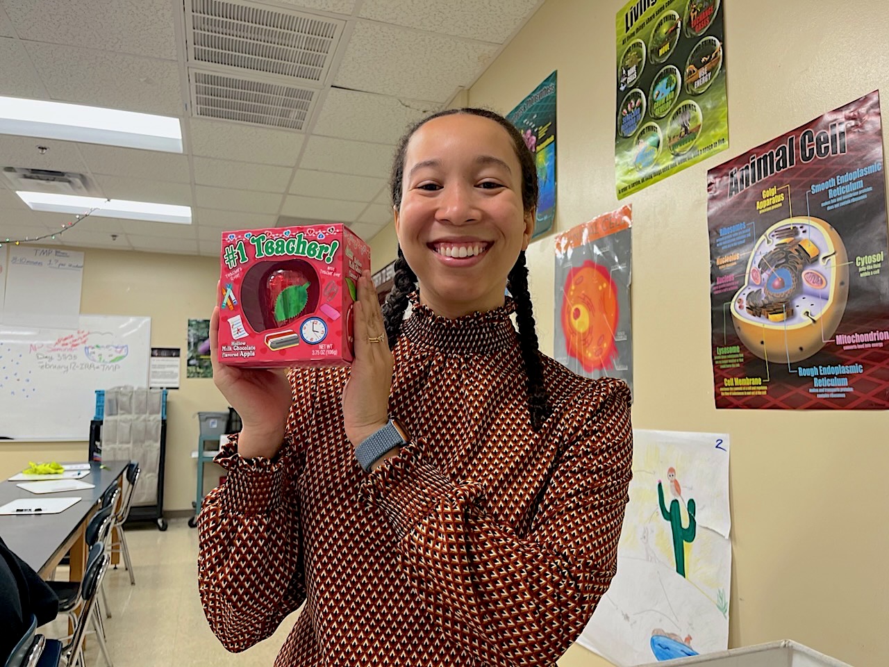 In a high school classroom, Hannah holds up a gift of a toy apple that says #1 teacher on the box, with science posters in the background.