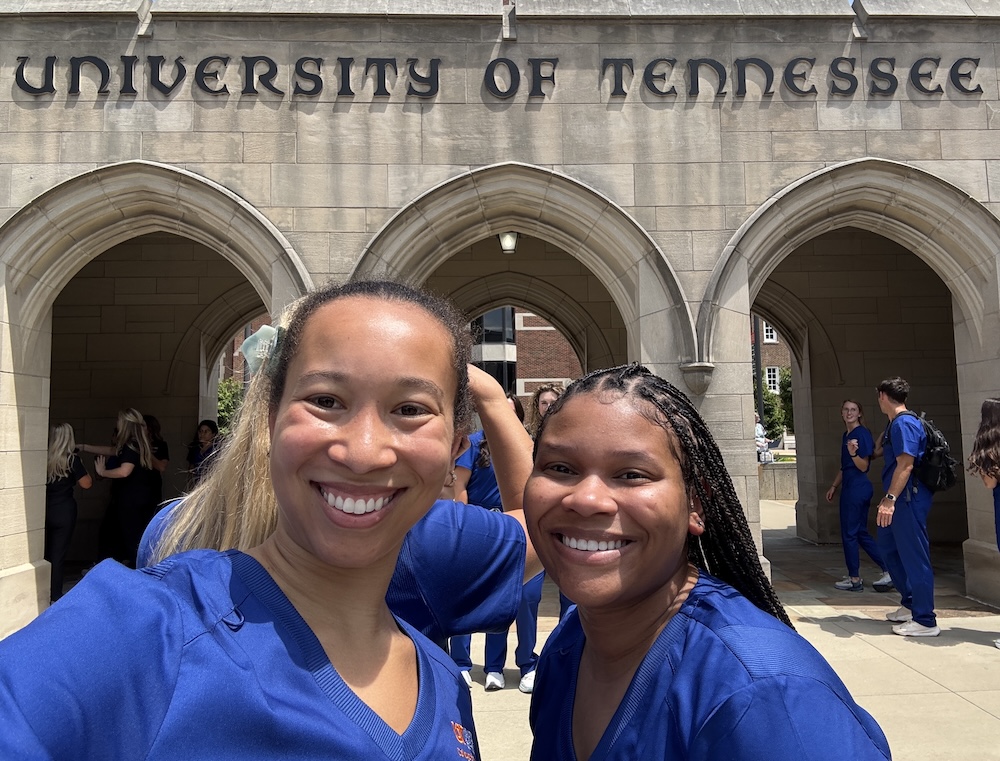 Hannah poses with a fellow Black student, both in blue scrubs, with others in the background and in front of the University of Tennessee sign over the campus arches in Memphis.