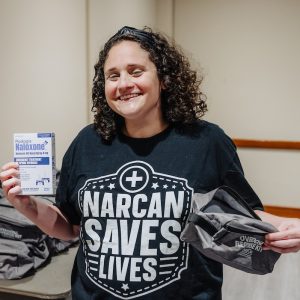 Dr. Jessi Gold is wearing a "Narcan Saves Lives" T-shirt, smiling, and holding a box of naloxone medicine and a gray bag it came in.
