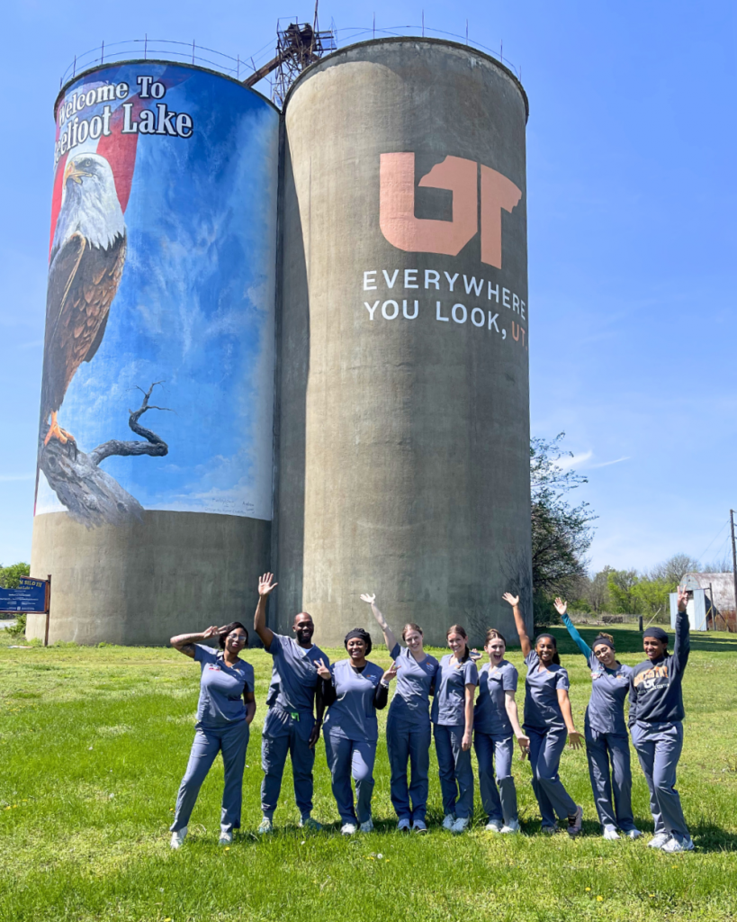 Students in gray scrubs are standing together, waving at the camera, in front of two huge silos, one with a bald eagle on it and one with the UT logo on it as well as the phrase "Everywhere you look, UT."