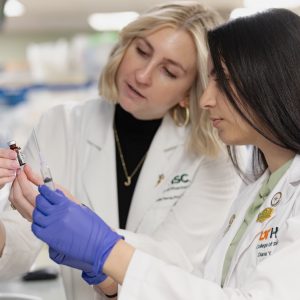 Two women wearing branded white coats examine a medicine vial and injection needle together.