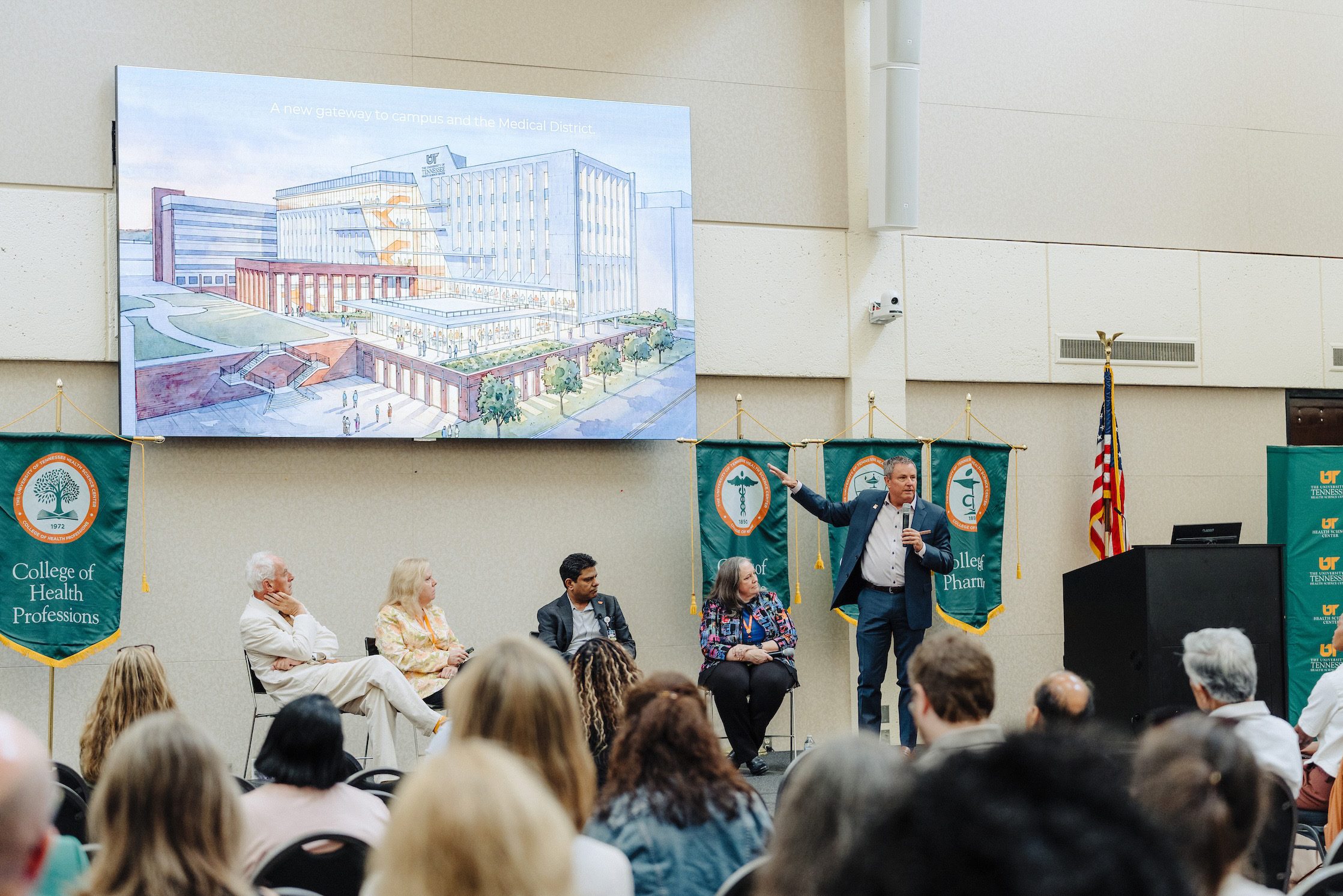 A wide shot from back of the seated audience shows Dr. Mike Hocker standing on stage gesturing toward a rendering of the College of Medicine Interdisciplinary Building projected on a screen above the four panelists.