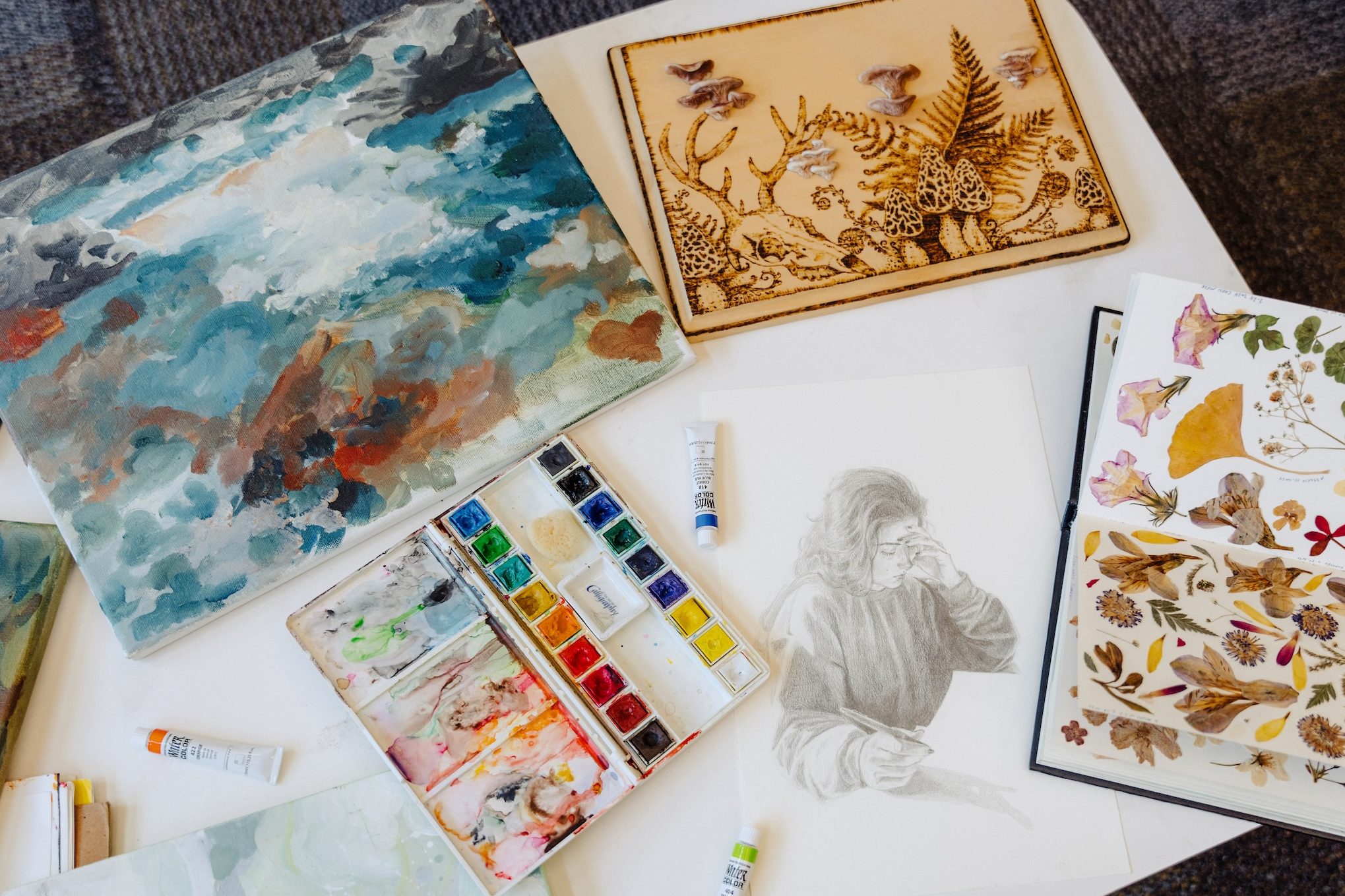 Art pieces, including paintings, illustrations, and collages of dried plants, displayed on a table.