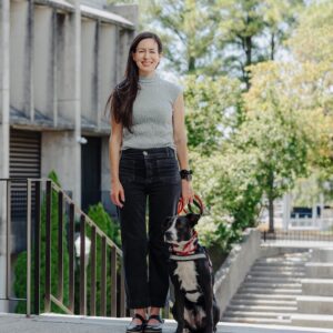 Dr. Corinne Webster and her dog outside the Student Alumni Center with green and white trees in the background.