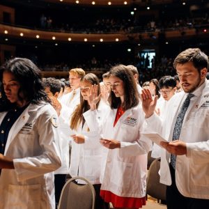 Inside a dim auditorium, students in short white coats hold up their right hands while reading an oath printed in a pamphlet.