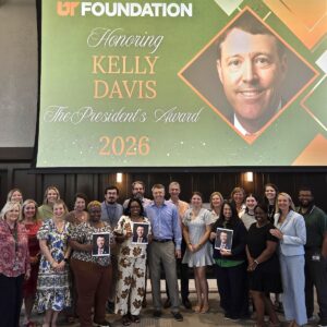 University Advancement team members pose with Kelly Davis beneath a presentation screen, the slide indicating his foundation President's Award. Some of the employees are holding headshots of Davis.